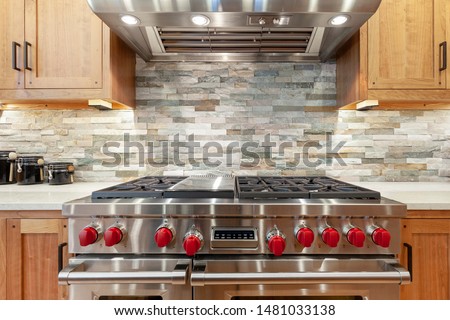 Stainless steel oven and stove with red knobs, stainless steel hood, brown kitchen cabinets, slate mosaic backsplash, natural stone, black jars Royalty-Free Stock Photo #1481033138