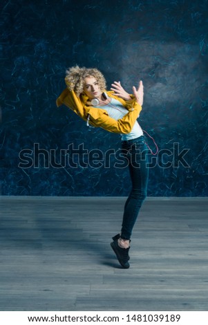 A handsome guy in a yellow jacket and headphones flies up in the dance, opens from the ground, his hand is bent in motion. On a dark background.	
