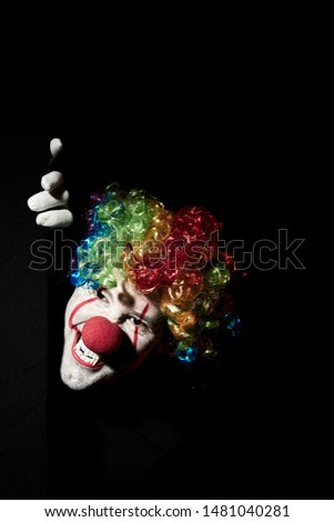 Scary clown peeping around the corner of a black wall. He is wearing a colored wig and sharp fangs.