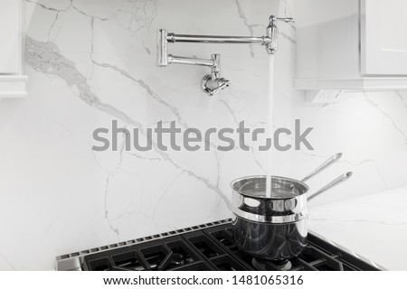 Granite backsplash with pot filler faucet, chrome pot on stove filling with water, white kitchen cabinets Royalty-Free Stock Photo #1481065316