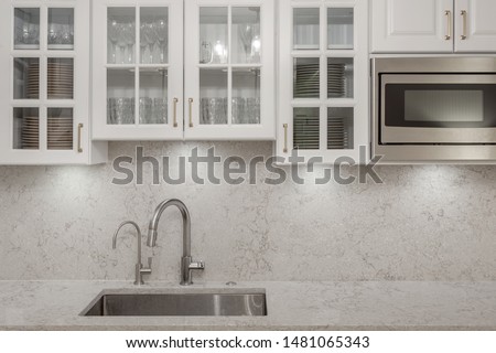 Glass door white kitchen cabinets with stainless steel microwave, kitchen faucet, soap dispenser, granite countertop and granite backsplash with gold color handles Royalty-Free Stock Photo #1481065343