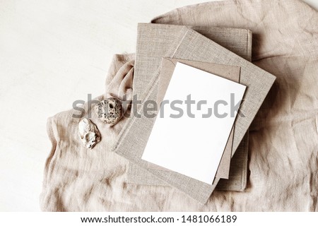 Feminine stationery, desktop mock-up scene. Blank greeting card, craft envelope, sea shells and old books on beige linen tablecloth background. Flat lay, top view. Summer rustic composition