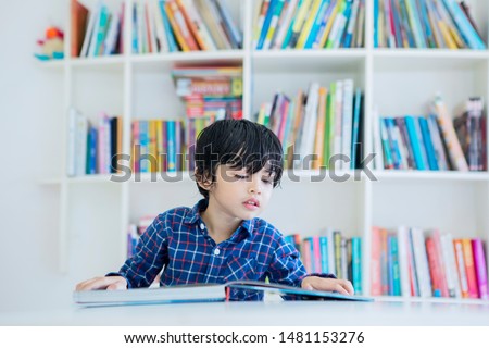 Picture of adorable little boy reading a book while studying in the library with bookcase background 