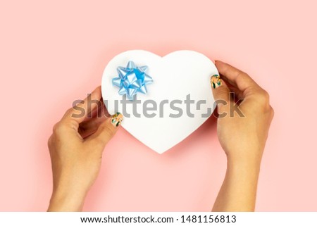 Female hands hold a shape heart red gift with a blue bow on a pink background. There is free space for text. Christmas, new year, valentine day concept.