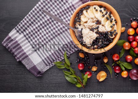 Picture of porridge with blueberries and banana slices in white plate on dark concrete background. Decoration of green leaves, aliches, plums and checkered cotton towels. Healthy breakfast concept.