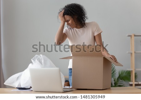 Disappointed african American millennial girl unpacking cardboard delivery box frustrated by delivered product, upset confused black young woman shopping online unwrap open bad quality product Royalty-Free Stock Photo #1481305949