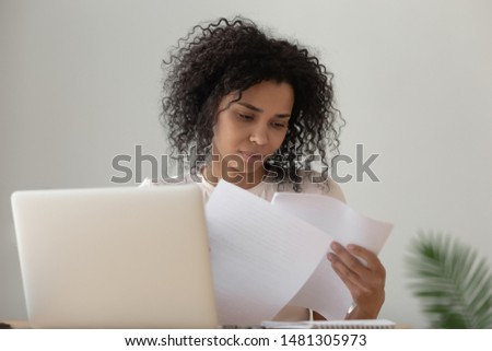 Concentrated biracial millennial girl sit at desk work on laptop reading paperwork education handouts, focused african American young woman busy studying on computer look through paper material Royalty-Free Stock Photo #1481305973