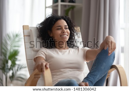 Happy peaceful african American millennial girl sit on chair relaxing at home or hotel dreaming or visualizing, smiling black young woman rest in rocker in living room look in distance thinking Royalty-Free Stock Photo #1481306042