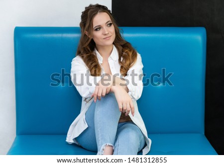 Photo waist-length portrait of a pretty brunette woman girl with long beautiful curly hair sitting on a sofa in a white shirt and blue jeans. Talking in different poses. Sitting facing the camera.