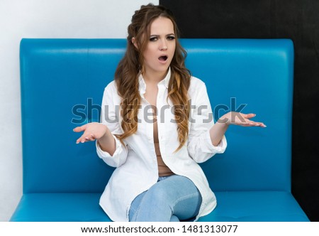 Photo waist-length portrait of a pretty brunette woman girl with long beautiful curly hair sitting on a sofa in a white shirt and blue jeans. Talking in different poses. Sitting facing the camera.