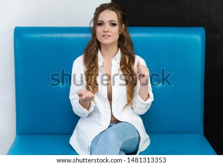 Photo waist-length portrait of a pretty brunette woman girl with long beautiful curly hair sitting on a sofa in a white shirt and blue jeans. Talking in different poses. Sitting facing the camera.