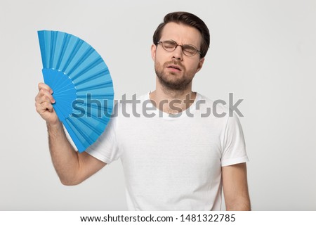Overheated millennial unhappy man in eyewear waving fan, suffering from high temperature inside or hot summer weather, isolated on grey white studio background. Young guy trying cool down, sweating. Royalty-Free Stock Photo #1481322785