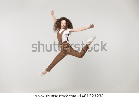 Happy young lady flying, jumping high with raised hands, feeling joyful, playful, having fun between softbox lights, posing for photo, celebrating victory, isolated on grey white studio background