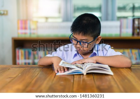 A cute Asian elementary school boy wearing blue glasses in a white school uniform is sitting, enjoying , reading comics in the library.