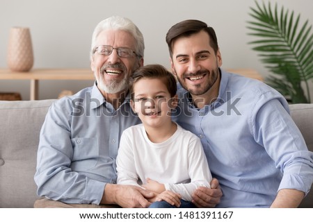 Different generation age caucasian men together at home, smiling grandpa grown up son and small cute grandson sitting on couch in living room looking at camera, multi generational male family concept Royalty-Free Stock Photo #1481391167