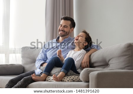 Little son sit on couch with father different age men laughing spend time watching comedy movie, cartoons feels happy, activities with kid at home, best friends, warm relations funny weekends concept