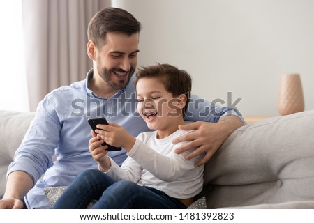 Young father embraces little son family sitting on couch at home using smart phone feels happy cheerful laughing on pranks online funny videos, parental control and modern tech everyday usage concept Royalty-Free Stock Photo #1481392823