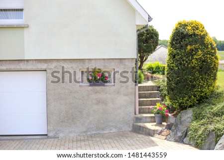 Village house with garden. Entrance to the garage in a country house. Geranium flowers on the window. Gray private house with a garden and flowers