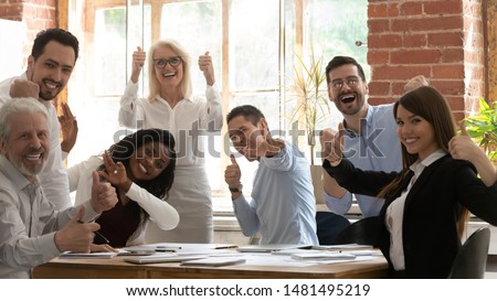 Excited multiracial businesspeople sit at office desk at meeting look at camera showing thumbs up together, overjoyed successful diverse colleagues posing for picture feel motivated at briefing
