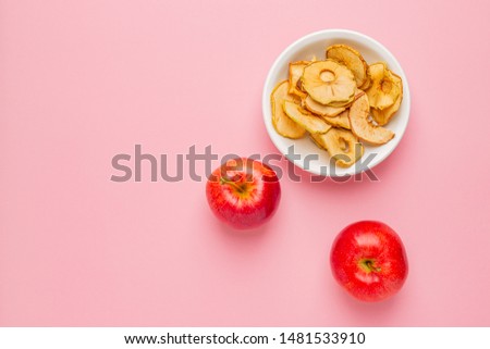 Dried apples chips in white ceramic bowl with fresh red apples on pink background