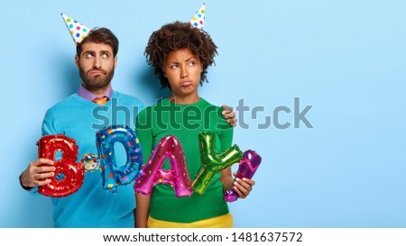 Photo of displeased mixed race couple embrace while hold colorful balloons meaning birthday, being in low spirit during celebration, make photo together, pose against blue studio wall. Copy space