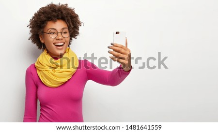 Lovely glad woman smiles broadly, takes selfie portrait on white cell phone, enjoys free time, makes pics for social networks, wears glasses, stylish yellow scarf and rosy jumper, smiles at camera