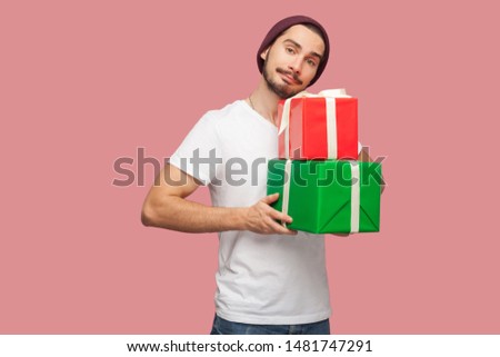 Portrait of handsome bearded young hipster man in white shirt and casual hat standing and holding two present box, looking at camera. Indoor, isolated, studio shot, pink background