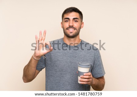 Handsome man with beard holding a take away coffee over isolated background showing ok sign with fingers