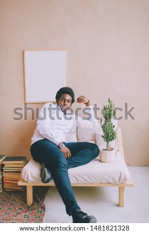 Lifestyle portrait of handsome young african black man sitting in chair  on textured wall with picture on background. Indoor portrait of happy dark skinned nigerian confident man relaxing at home