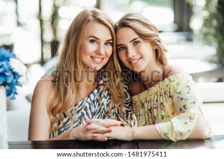 A young beautiful blonde girl and her pretty mom rest on a summer terrace cafe, communicate. They are happy and funny.