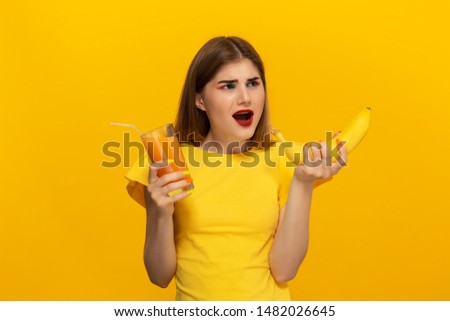 Close-up portrait of a beautiful young girl holding glass of ofange juice and banana close to face isolated over yellow background. Concept of beauty and health care.