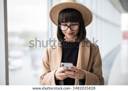 Young woman wear in coat and hat with glasses talking on mobile phone while standing by window in office.