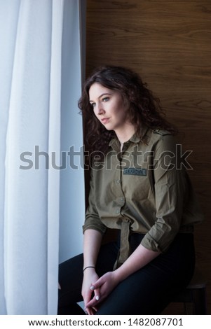Young woman sitting next to a window, looking pensive through curtains.
