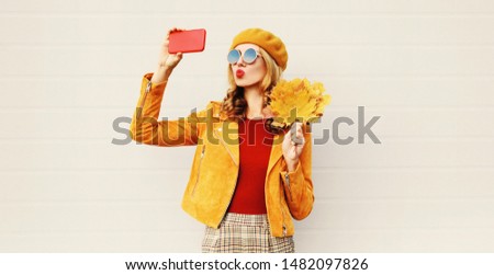 Autumn mood! woman taking selfie picture by phone blowing red lips sending sweet air kiss holding yellow maple leaves wearing french beret hat posing on city street over gray wall background