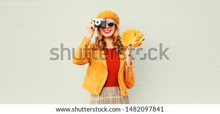 happy smiling woman with autumn yellow maple leaves, retro camera taking picture in french beret over gray wall background