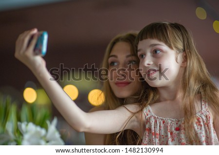 Girl taking a selfie with her mother or sister. Little and adult sisters take a selfie. A child and an adult girl with brown hair. Girls in the cafe take pictures and look at the smartphone.Boke