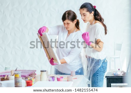 Creative woman painter teaching female student creating fluid acrylic abstract painting in art therapy class, with table full of bottle with defferent paint colors. Art, design, creating concept.