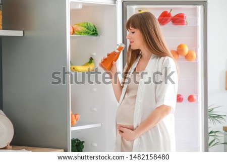 Beautiful pregnant woman drinking juice near fridge in kitchen