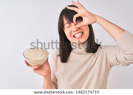 Young beautiful Chinese woman holding bowl with rice over isolated white background with happy face smiling doing ok sign with hand on eye looking through fingers