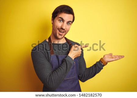 Young handsome shopkeeper man wearing apron standing over isolated yellow background amazed and smiling to the camera while presenting with hand and pointing with finger.