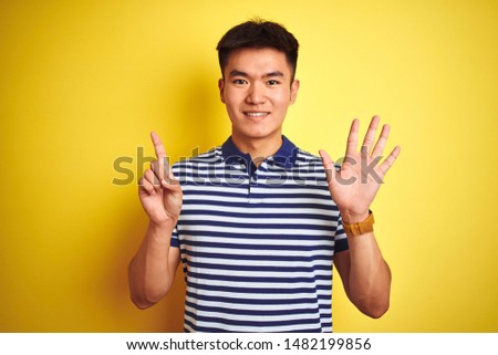 Young asian chinese man wearing striped polo standing over isolated yellow background showing and pointing up with fingers number six while smiling confident and happy.