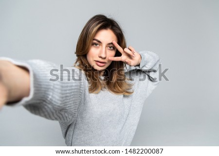 Pretty cheerful brunette woman showing peace gesture while taking selfie on smartphone over gray background