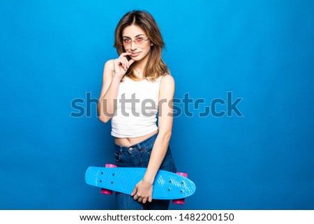 Portrait of pretty young woman in vivid casual clothes standing, looking camera, holding yellow skateboard isolated on blue wall background in studio.