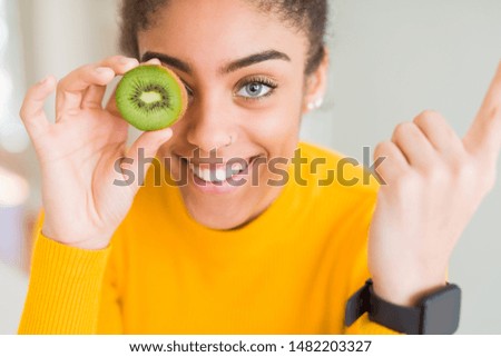 Young african american girl eating green kiwi very happy pointing with hand and finger to the side