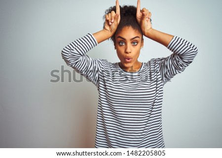 African american woman wearing navy striped t-shirt standing over isolated white background doing funny gesture with finger over head as bull horns