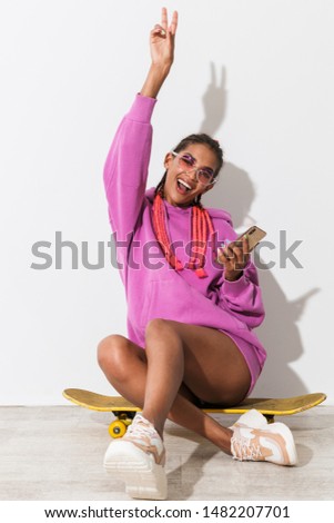 Picture of happy emotional young african woman isolated over white wall background in bright pink sweatshirt sit on skateboard using mobile phone.