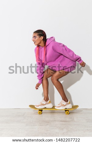 Photo of gorgeous emotional happy young african woman skater isolated over white wall background in bright pink sweatshirt on skateboard.