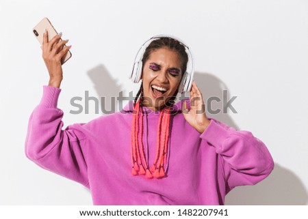 Picture of happy screaming singing young african woman isolated over white wall background in bright pink sweatshirt using mobile phone listening music with headphones.