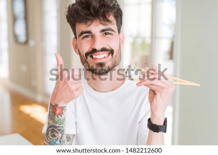 Young man eating asian sushi using chopsticks happy with big smile doing ok sign, thumb up with fingers, excellent sign
