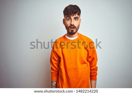 Young man with tattoo wearing orange sweater standing over isolated white background afraid and shocked with surprise expression, fear and excited face.